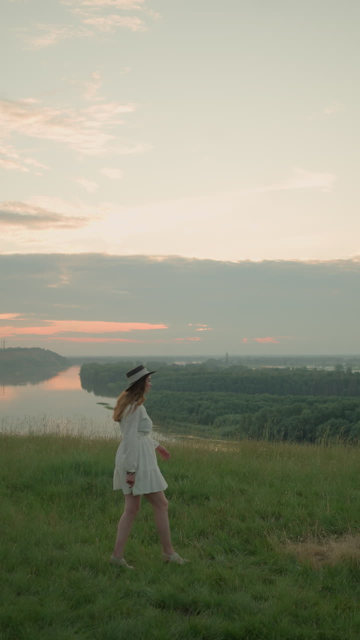 색 드레스와 모자를 입은 한 여인이 해가 지는 동안 잔디을 가로질러 평온한 호수를 향해 우아하게 걸어가고 있다.그녀는 잠시 멈추고, 귀에 손을 고, 평온하고 내성적인 순간을 만니다.