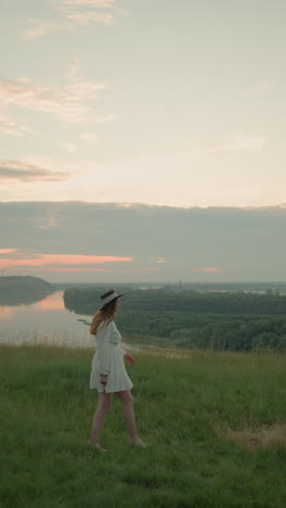una mujer con un vestido blanco y un sombrero camina con gracia a través de un campo cubierto de hierba hacia un lago sereno al atardecer. se detiene, colocando sus manos sobre sus orejas, creando un momento tranquilo e introspectivo