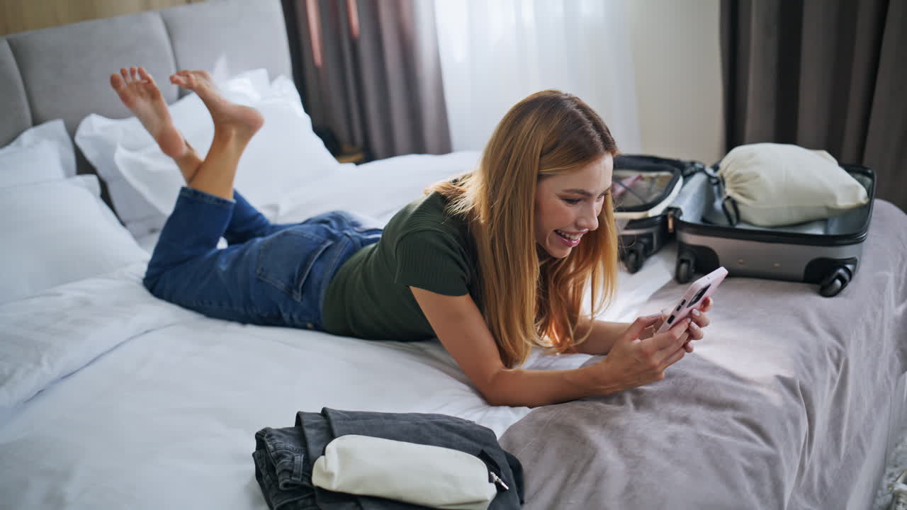 Cheerful girl texting hotel bed arriving on journey. Smiling woman messaging