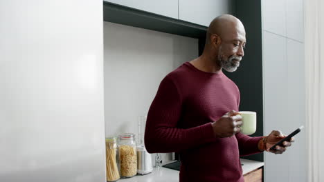 happy biracial man drinking coffee and using smartphone in kitchen, slow motion