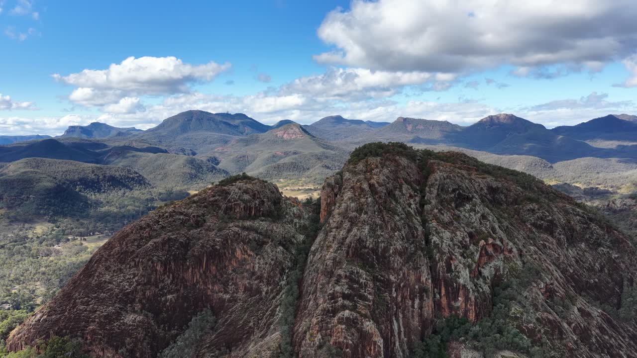 Drone camera rises above Split Rock, revealing rugged sandstone formations, dense forest, and expansive mountain views under bright daylight with scattered clouds