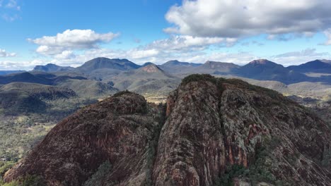 drone camera rises above split rock, revealing rugged sandstone formations, dense forest, and expansive mountain views under bright daylight with scattered clouds