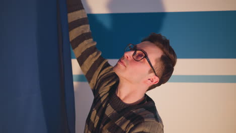 male technician adjusts blue studio curtain during broadcast set prep under warm spotlight, striped backdrop ladder visible behind, cables trailing floor, rigging and camera equipment visible