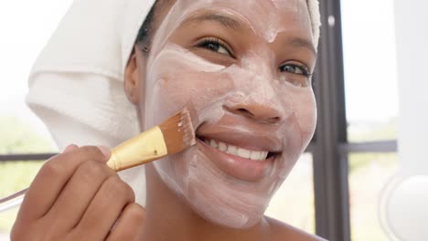 Portrait-of-happy-biracial-woman-with-towel-on-head-making-mask-on-face-in-bathroom,-slow-motion