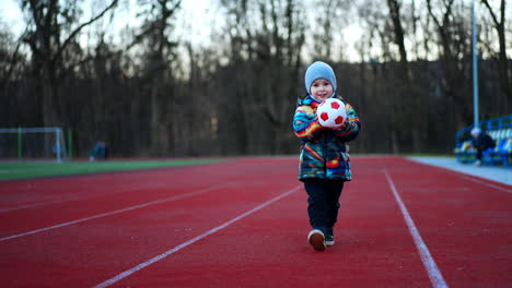 cute kid approaches the camera carrying a ball. lovely smiling baby having fun at the stadium.