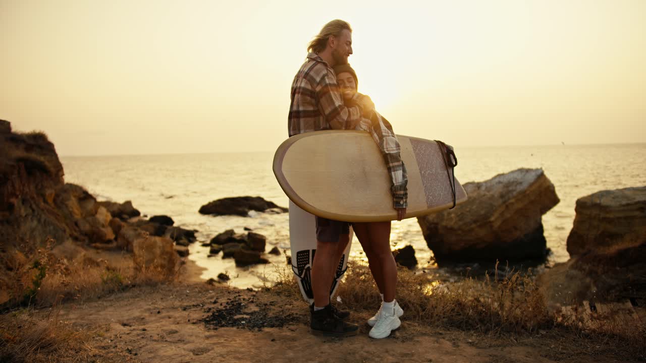Romantic meeting of a blond guy with a beard and his blonde girlfriend in a checkered shirt, who holds a surfboard and hugs her boyfriend on a rocky shore near the sea