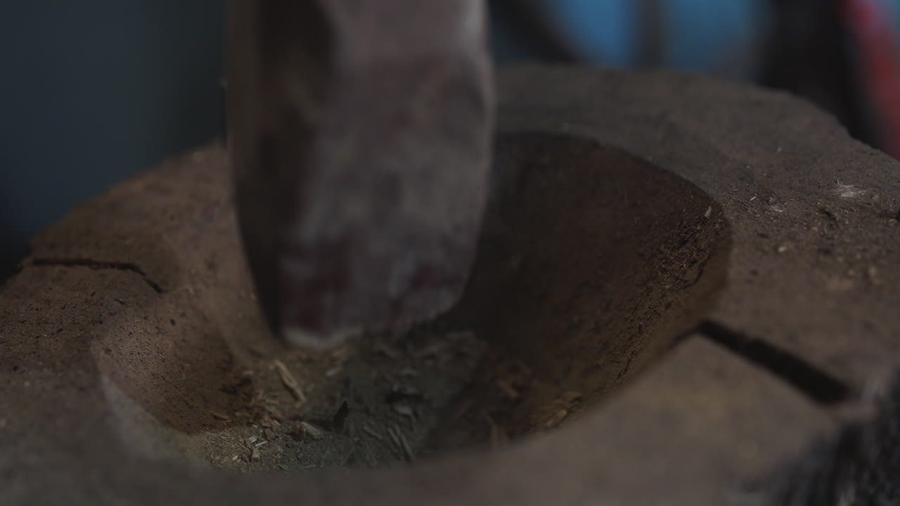 Heavy wooden pestle crushes Ilex paraguariensis leaves inside rustic mortar during traditional yerba mate grinding process, close-up of tool impact inside hollowed grinding cavity, slow motion