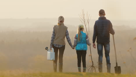 Familia-Con-Una-Plántula-De-árbol,-Un-Aspersor-Y-Una-Pala-De-Pie-En-Un-Lugar-Pintoresco