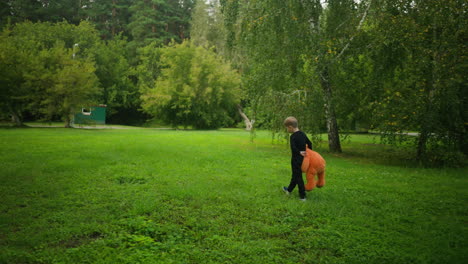 rear view of young boy walking through green grassy area carrying large orange teddy bear, heading toward white fence with trees and official-style building in background under daylight sky