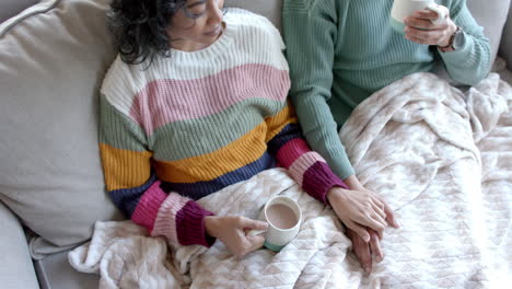 una feliz pareja biracial sentada en el sofá y bebiendo café bajo una manta en casa, en cámara lenta