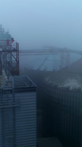 tops of the elevator tanks hardly seen in grey fog and thick white smoke coming from pipes. aerial view on the modern granary complex plant. vertical video