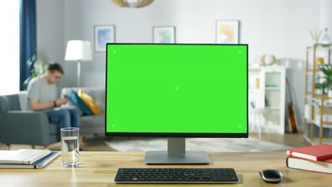 una computadora personal moderna con una pantalla de maqueta verde de pie en el escritorio de la acogedora sala de estar. un hombre con un teléfono móvil se relaja en su silla.