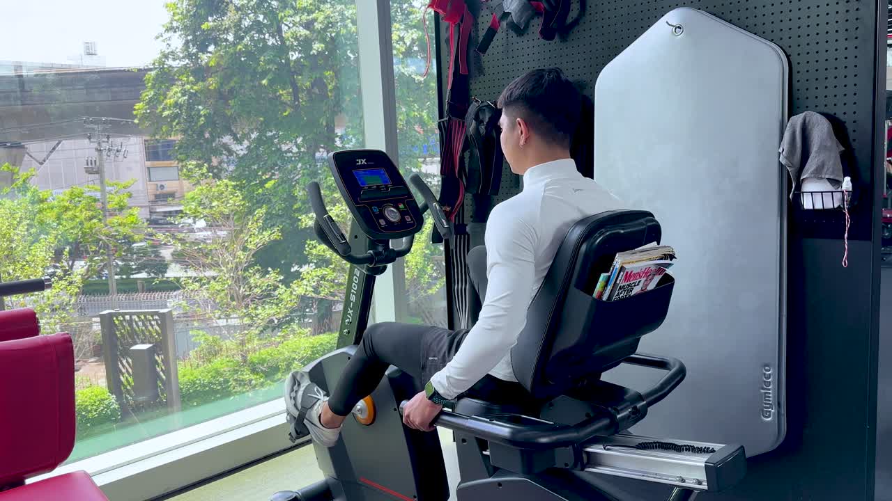 Young man exercises on recumbent bike in bright gym, natural daylight, side view, steady shot
