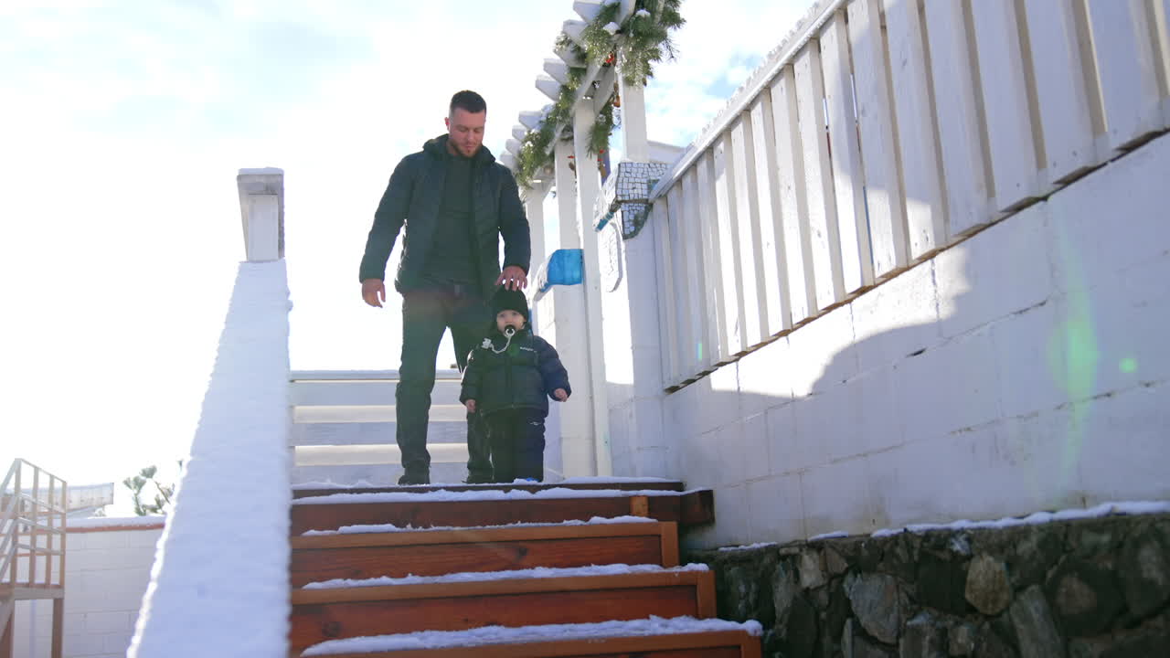 Man and a toddler boy wearing warm clothes stand on the stairs. Father and his little son with pacifier in mouth outdoors in winter. Low angle view.