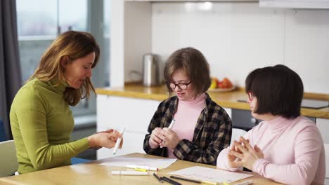 Zwei-Mädchen-Mit-Syndrom-Zeichnen-Gemeinsam-In-Der-Küche-Neben-Der-Lehrerin-Oder-Mutter,-Zeitlupe