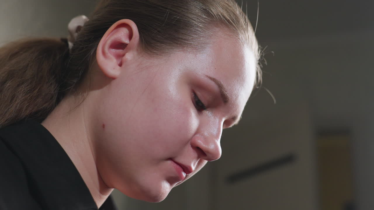 Side close up of teenager indoors, face angled downward, eyes intent on task, calm focus under soft room light, natural complexion visible, patient attention suggesting careful work off frame