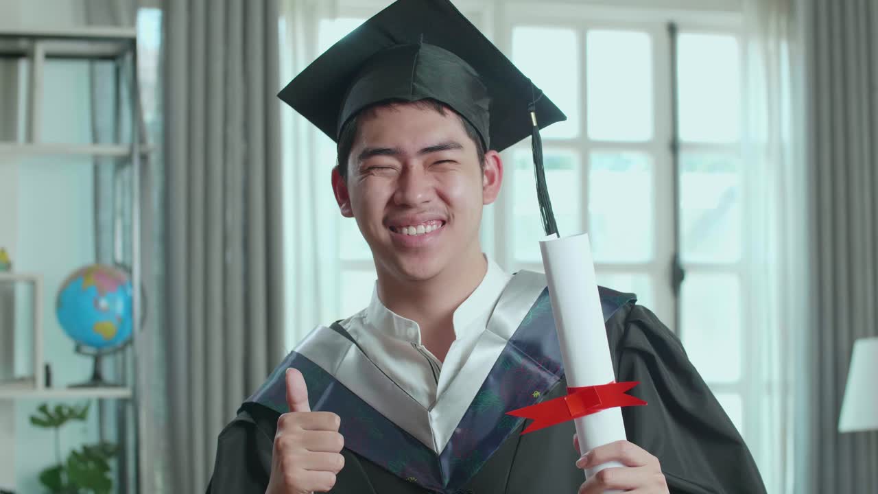 hombre asiático emocionado sosteniendo un certificado universitario, sonriendo y el pulgar hacia la cámara. graduado masculino con un vestido de graduación y gorra sentado en la sala de estar