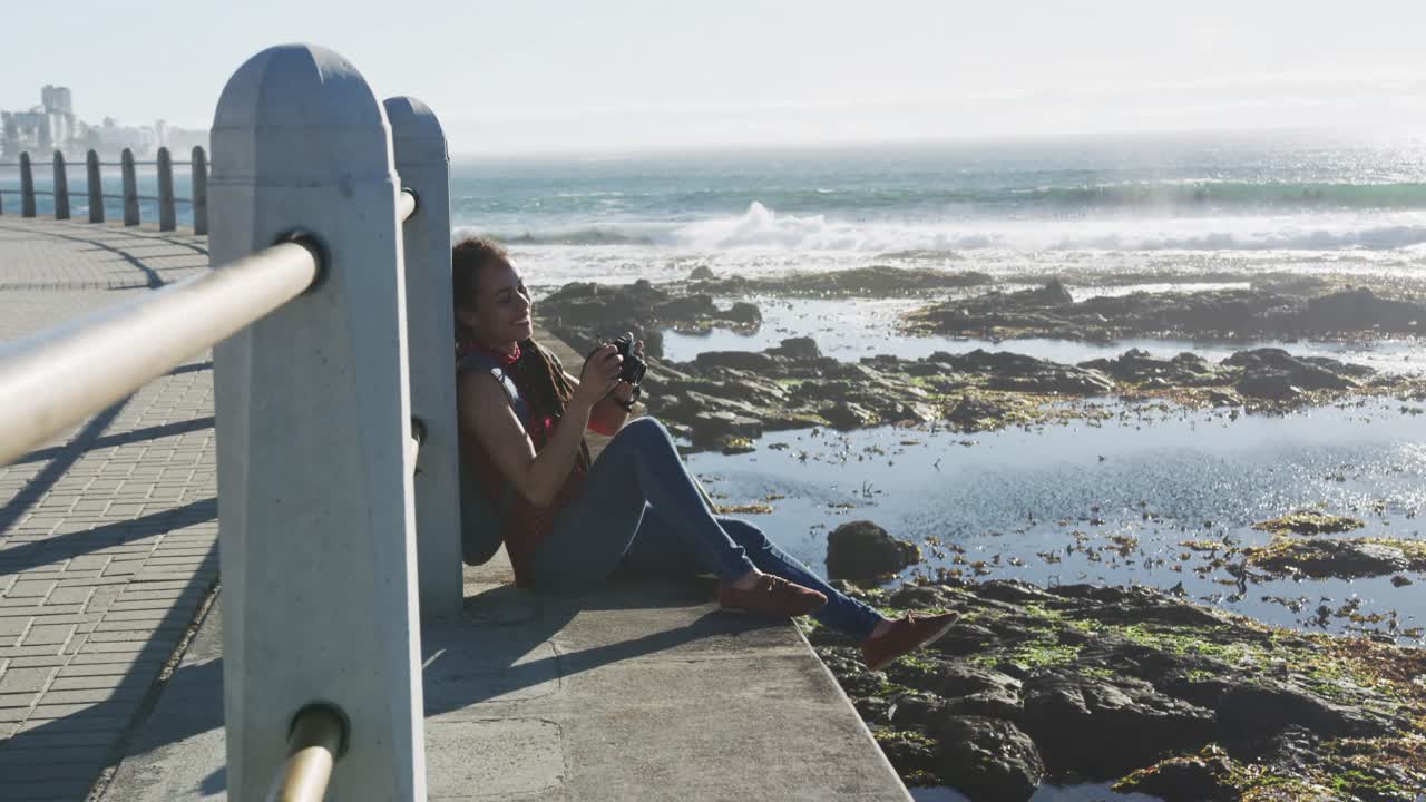 African american woman sitting and photographing on promenade by the sea