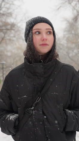 girl in black hoodie carrying guitar in pack looks around thoughtfully while snow falls gently, with a snow-covered bench in the background enhancing the wintry scene