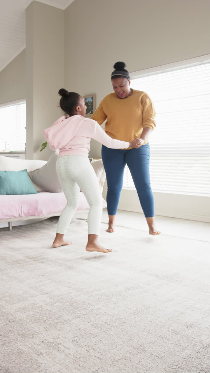 Vertical video of happy african american mother and daughter dancing in living room, in slow motion