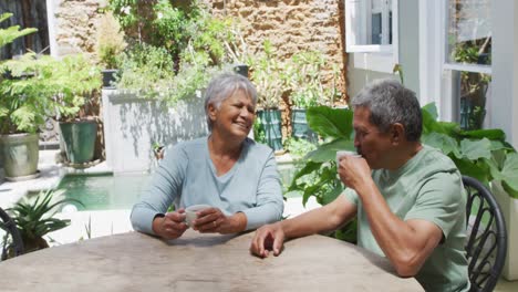 Happy-senior-mixed-race-couple-sitting-in-garden-drinking-coffee