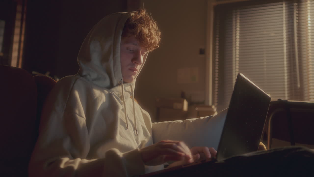 Young Man in Hoodie Coding on Laptop in Softly Lit Room