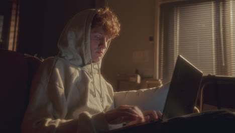 Young Man in Hoodie Coding on Laptop in Softly Lit Room