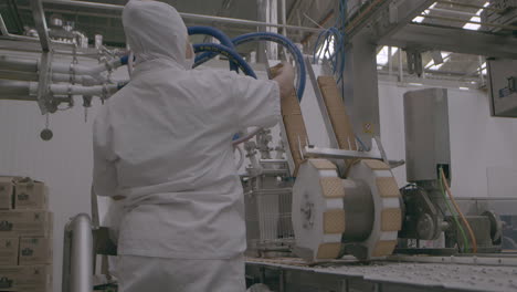 Factory-worker-in-protective-equipment-operating-machinary-by-loading-biscuits-onto-wheel,-which-assembles-ice-cream-sandwiches