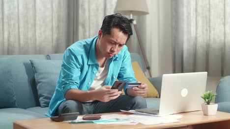 hombre asiático con una computadora portátil con tarjeta de crédito y teléfono inteligente sacudiendo la cabeza debido a no poder pagar dinero