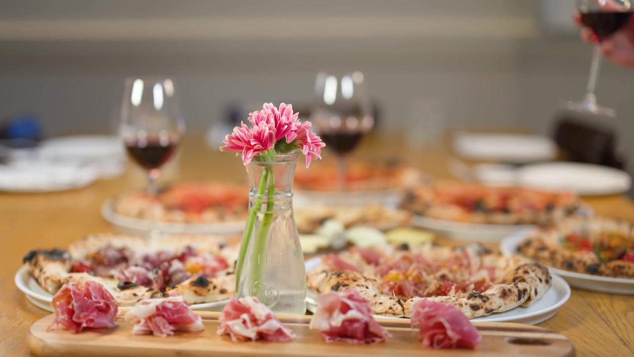 Inquadratura media statica di un tavolo da pranzo italiano con pizza, prosciutto, vino e fiori rosa