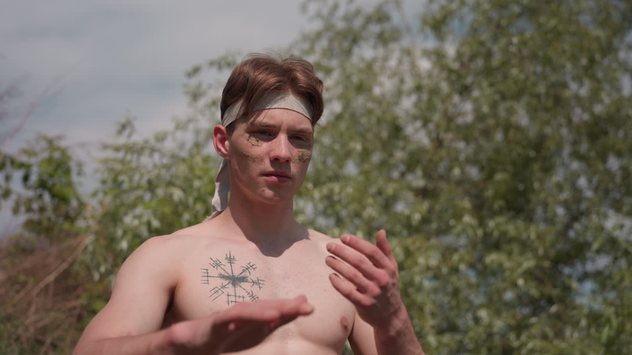 muscular military man loosening fabric headband with both hands pushing head back in relief after intense outdoor training session by riverbank dirt and grass under cloudy blue sky setting