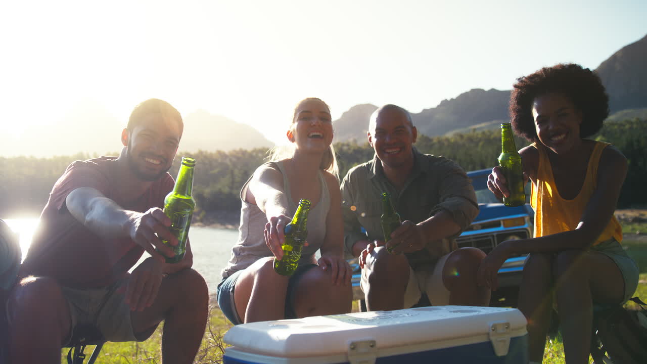 Ritratto di amici con zaini accanto a un pick-up, in viaggio, che bevono birra da un frigo portatile in riva al lago