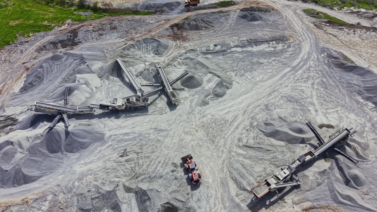 Granite quarry operation is active with large machinery and stone piles under bright sunlight in the afternoon