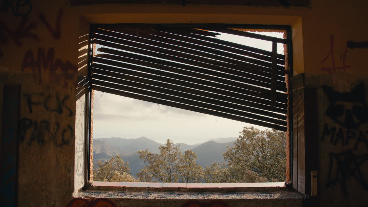 Sunlight filters through tilted wooden slats of a broken window, revealing a serene mountain view framed by an abandoned, graffiti-covered wall