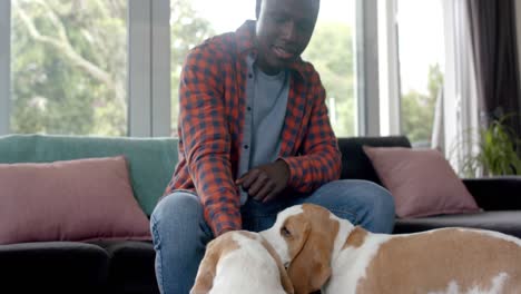 Happy-african-american-man-sitting-on-sofa,-petting-his-pet-dogs-at-home,-slow-motion