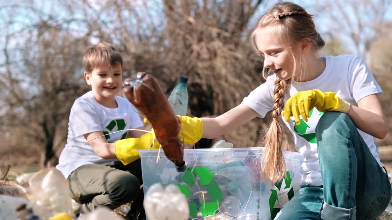 Boy and girl collecting plastic garbage in a container in a polluted clearing, recycling signs on the T-shirts. Slow motion