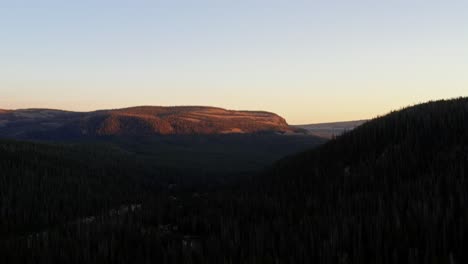 Impresionante-Toma-Aérea-Del-Paisaje-De-Un-Cañón-En-El-Bosque-Nacional-Uinta-Wasatch-Cache-En-Utah-Con-Una-Carretera-Debajo-Rodeada-De-Pinos-Y-Montañas-Rocosas-Durante-Una-Vívida-Puesta-De-Sol-De-Verano