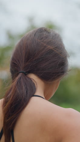 back view of woman stretching neck gently in park with trees in blurred background, capturing peaceful outdoor stretching and neck flexibility in a natural setting