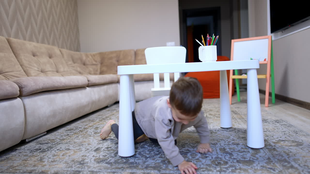 Happy Caucasian toddler crawls by the floor under the desk. Portrait of an energetic playful baby boy playing at home.