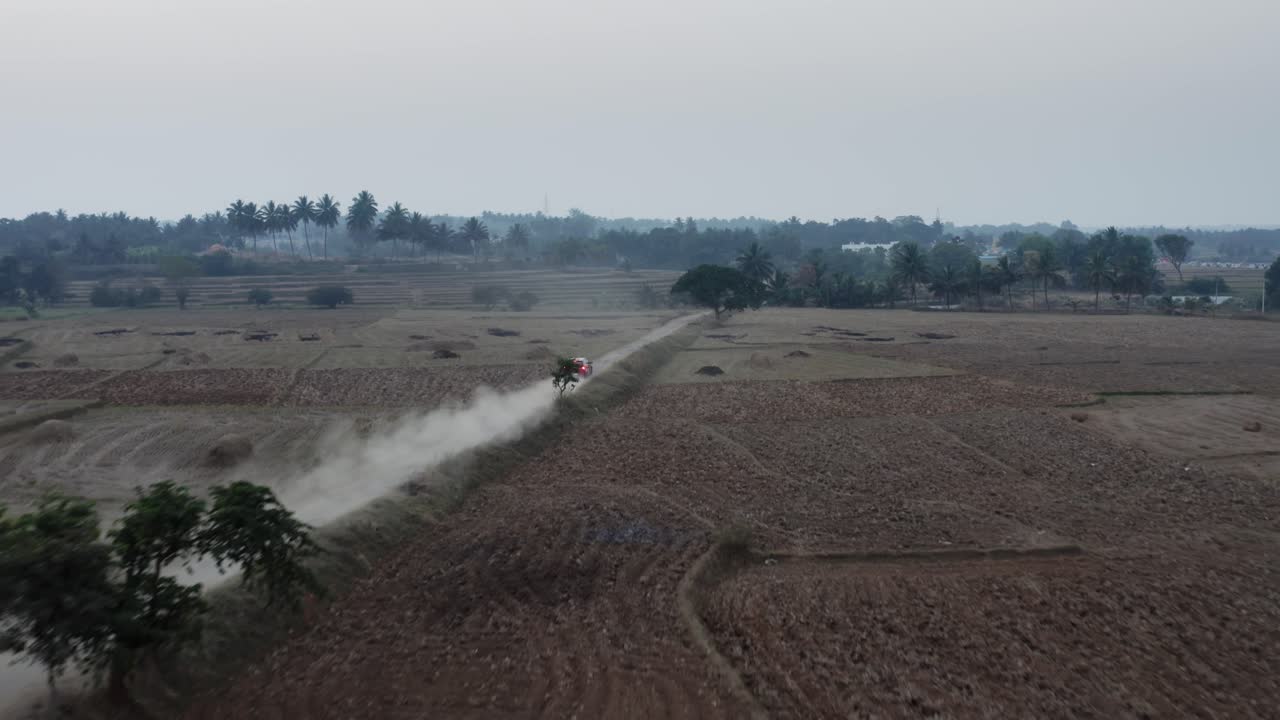 Um veículo vermelho acelera por uma estrada de terra estreita atravessando campos abertos, deixando um rastro de poeira para trás, enquanto palmeiras e uma vila distante delineiam o horizonte