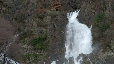 Tall-powerful-waterfall-on-rocky-autumn-forest-hill,-Pan-right