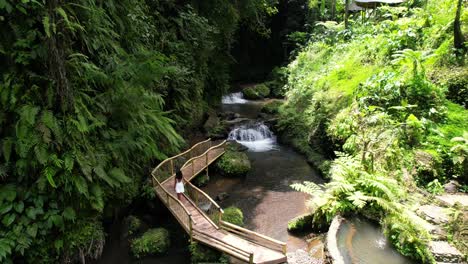 mujer vestida de blanco camina sobre un puente de bambú a lo largo de un río y una cascada en la jungla de bali indonesia