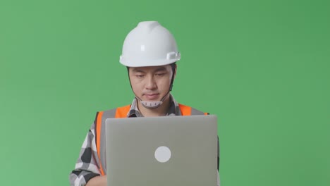 primer plano de un ingeniero masculino asiático con casco de seguridad trabajando en una computadora portátil mientras camina en el estudio de fondo de la pantalla verde
