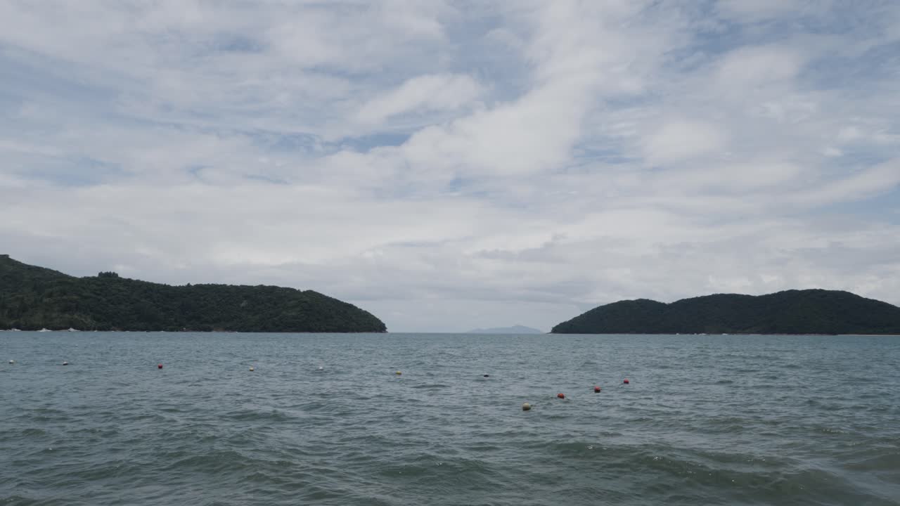 Dramatic clouds mostly-cover a blue sky above two south Atlantic ocean islands, connected by green-gray waves of seawater.
