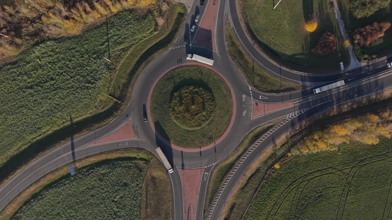 Aerial view of a roundabout surrounded by green fields and intersecting roads. Cars navigate the circular traffic pattern under daylight, showcasing urban infrastructure and transportation flow