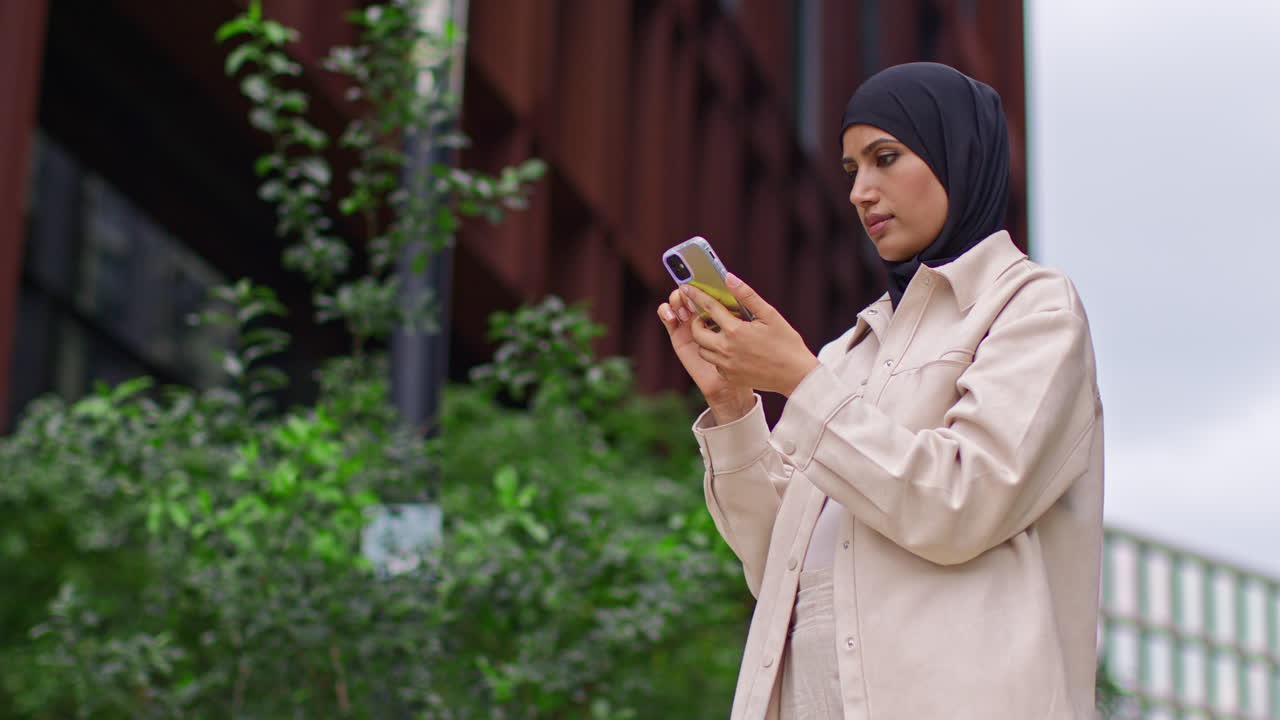 Muslim Businesswoman Wearing Hijab Going To Work Standing Outside Modern Office Looking At Mobile Phone 7
