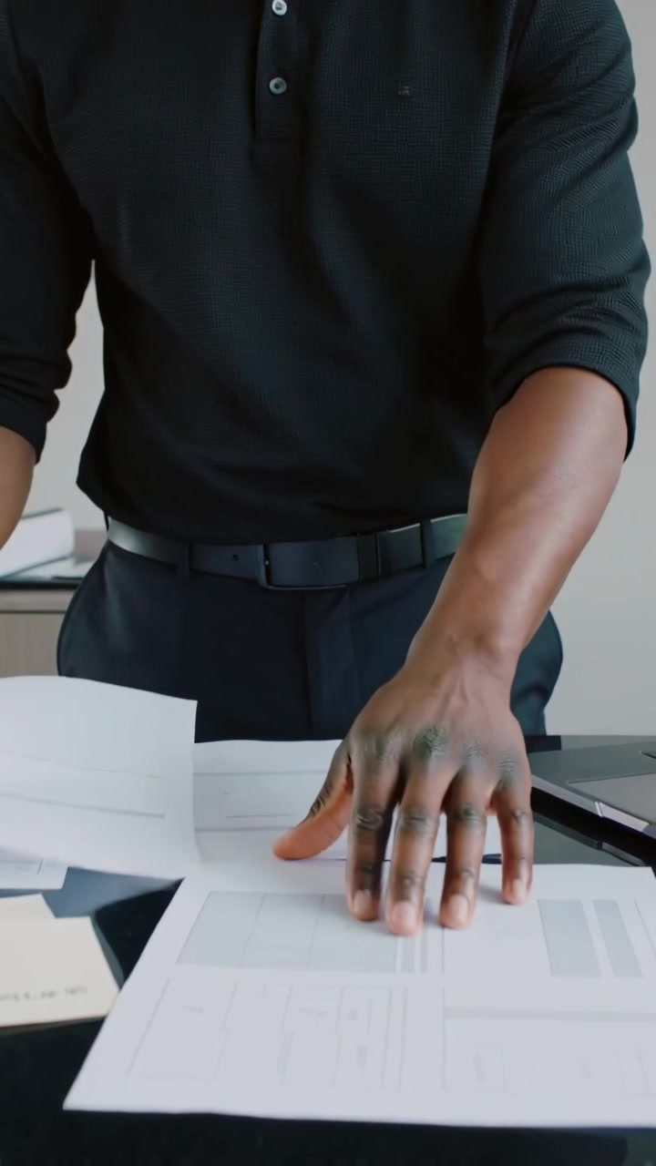 Vertical video: Holding sheet, man in polo pointing and reviewing papers at office desk with laptop