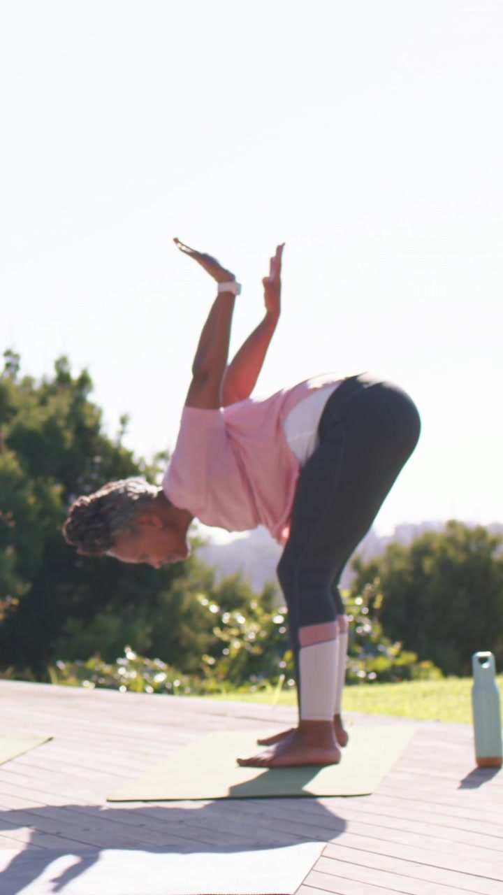Vertical video: Folding woman from raised arms, rising for balance on mat on deck, wearing pink top