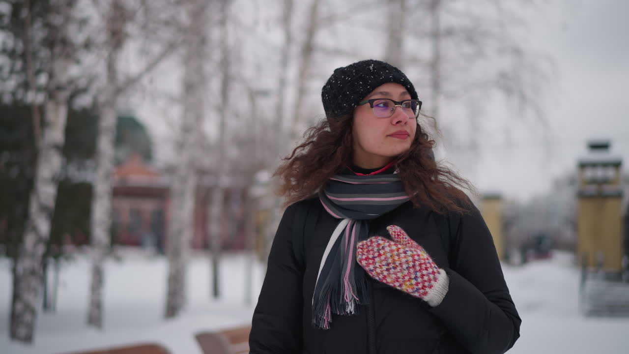 mujer de pie en un parque nevado de la ciudad en un día de invierno, con abrigo negro, bufanda, gorro, gafas, pelo rizado, en el exterior, soledad, estación fría, ambiente gélido, entorno congelado, fondo tranquilo, cielo cubierto, estilo de vida tranquilo