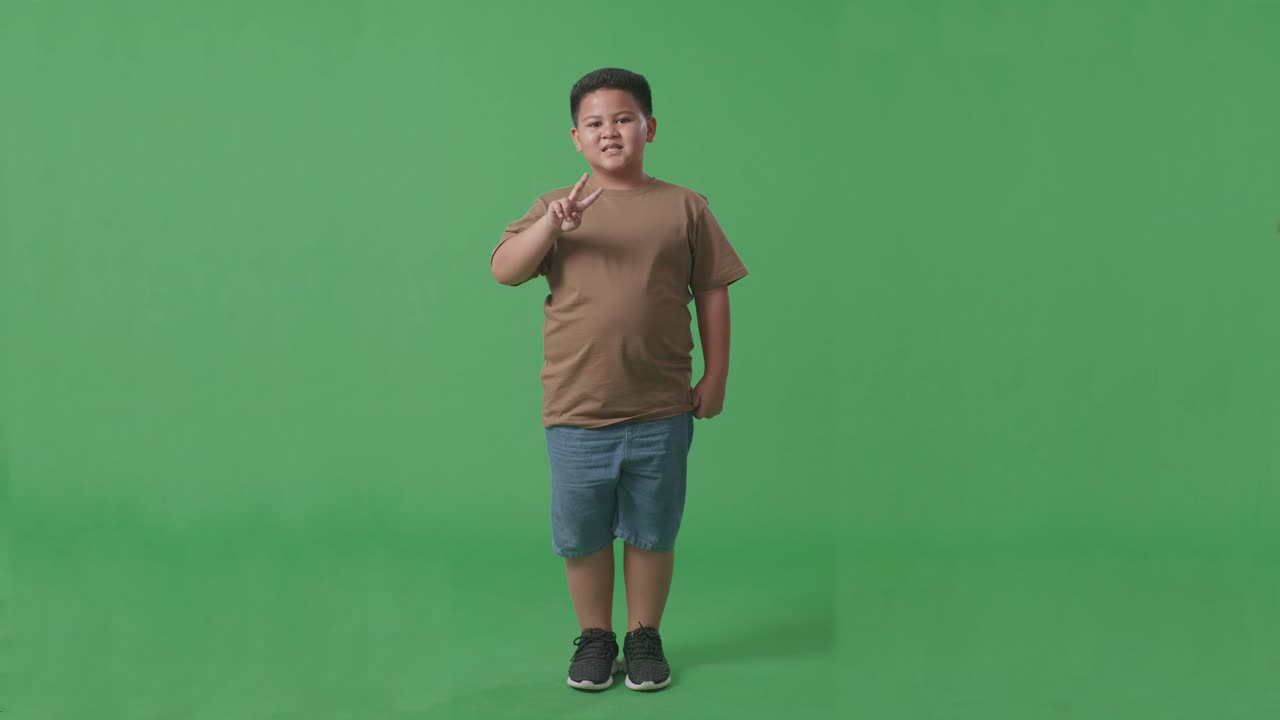 cuerpo lleno de niño asiático mostrando gesto paz y sonriendo mientras estaba de pie en la pantalla verde en el estudio